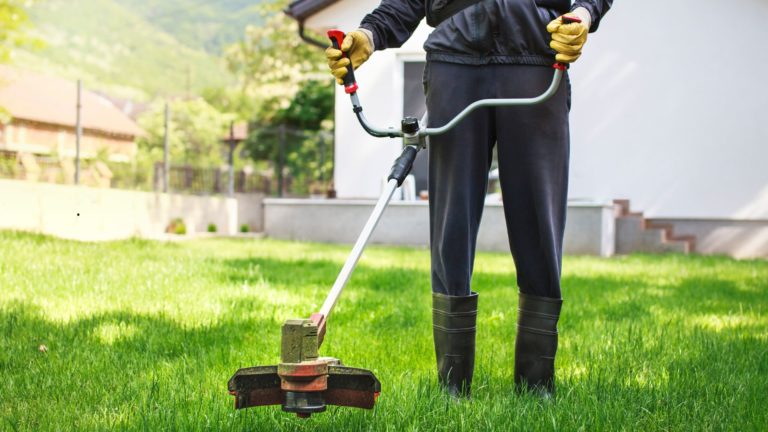 Persona che utilizza un decespugliatore su un prato verde, con guanti di sicurezza e stivali di gomma