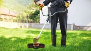 Persona che utilizza un decespugliatore su un prato verde, con guanti di sicurezza e stivali di gomma
